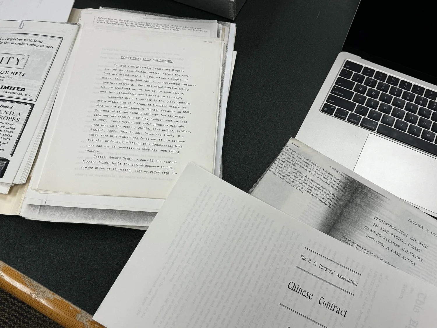 Archival documents about salmon canning and Chinese contract labour arranged beside a laptop.
